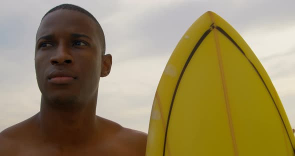 Front view of African American male surfer standing with surfboard on the beach 4k