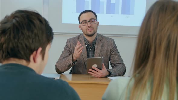 Male Teacher Holding a Tablet Sitting in Front of Class