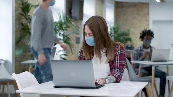 Confident Young Woman in Covid Face Mask Typing on Laptop Keyboard As Redhead Caucasian Man Walking