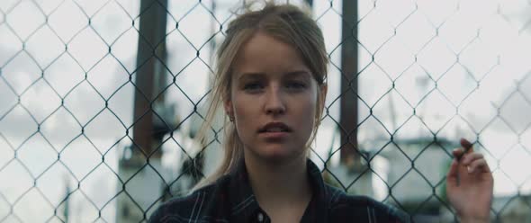 Young woman looks into the camera in front of metal fence and industrial blurry background