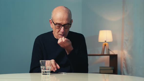 Elderly Man with Eyeglasses Sitting at the Table at Home Eats a Heart Pill