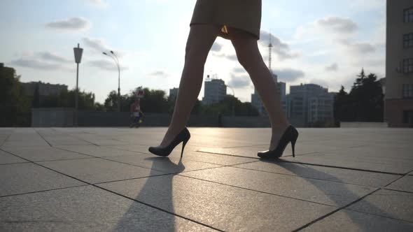 Slim Female Legs in Black Shoes on High Heels Walking on City Square at Sunset