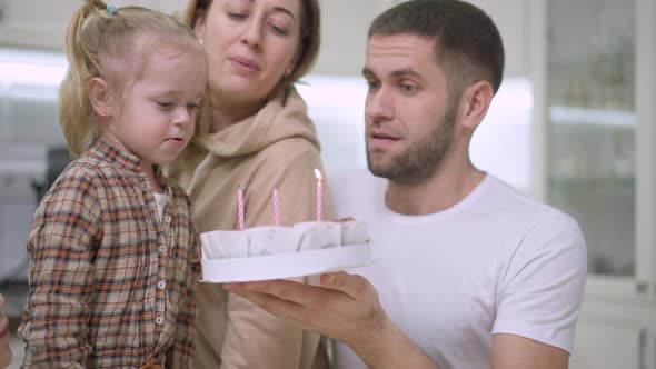 Cute Caucasian Baby Girl Blowing Candles on Birthday Cake with Blurred Parents Helping at Background