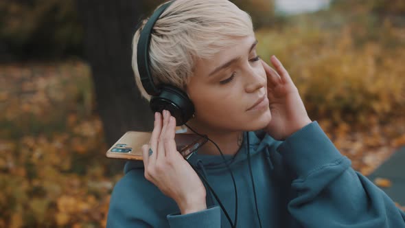 Young Blond Woman Listening To the Music From Her Smartphone in the Park Using the Headphones