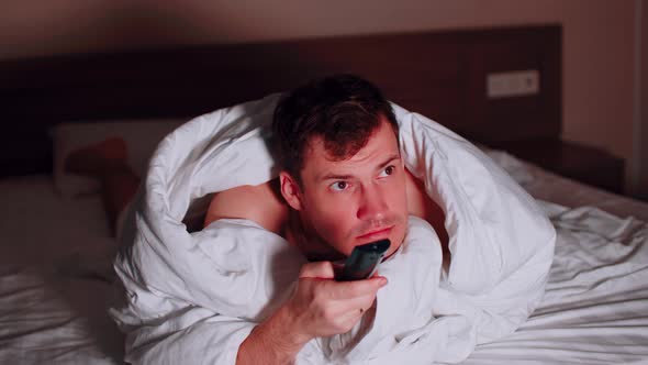 Young Man Under Blanket Lying on Bed Watching Tv and Waving His Legs in Late Evening