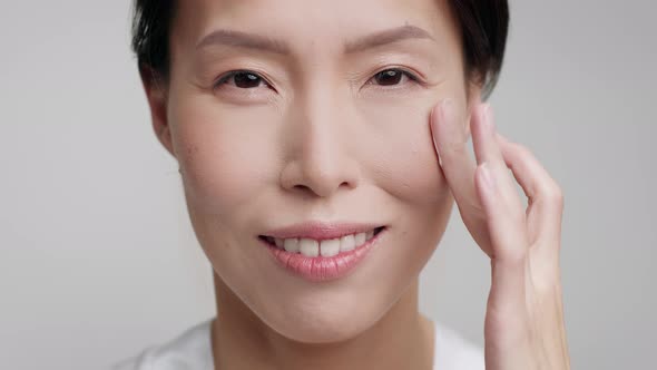 Mature Chinese Lady Applying Moisturizer Cream On Face Gray Background