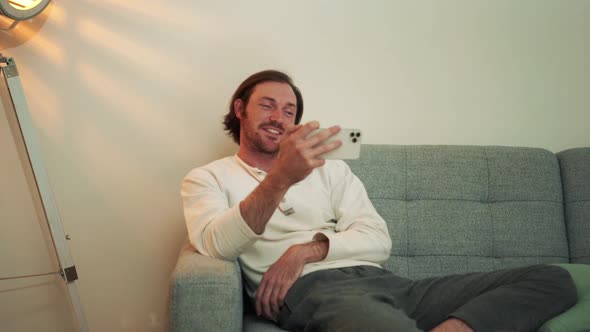 Positive long-haired man talking by video call on phone while sitting on the sofa