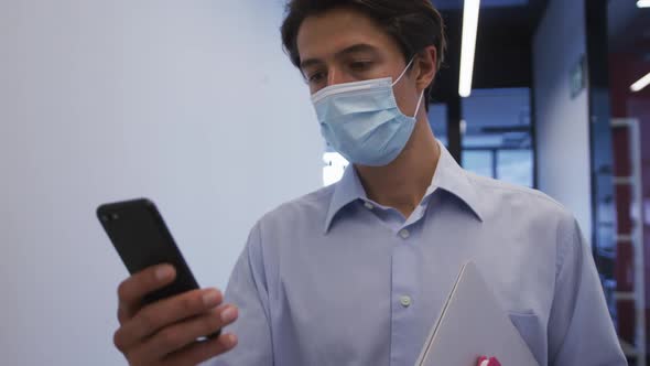 Caucasian businessman wearing face mask walking using smartphone in office corridor