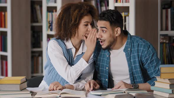 Two Students of Classmate Sitting at Desk in Lesson Doing Homework in Library Girl Whispers in Ear