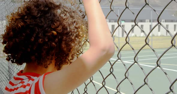 Rear view of mixed-race schoolgirl standing near wire mesh fence at school 4k