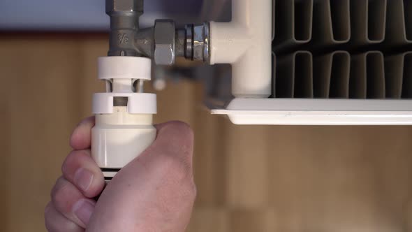 A man s hand turns a thermostat on a white heating radiator