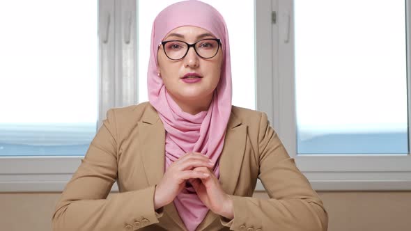 Woman in Hijab and Glasses Sits at the Table and Talks to the Camera Zooming Out of the Camera