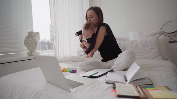 Young Mother Freelancer with Baby Son Sitting on Bed and Trying to Work at Laptop