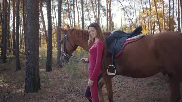 Side View of a Young Caucasian Girl in Pink Clothes Walking with Beautiful Brown Horse Between Trees