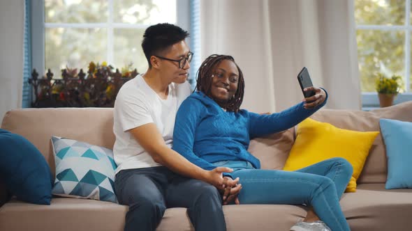 Young Diverse Joyful Couple Hugging and Smiling While Taking Selfie Photo in Living Room