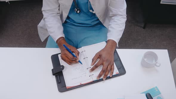 Black Male Doctor Writes Notes Sitting at the Desk Wearing Medical Gown