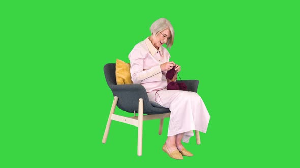 Senior Lady Sitting in an Armchair and Knitting and Looking To Camera at Some Moment on a Green