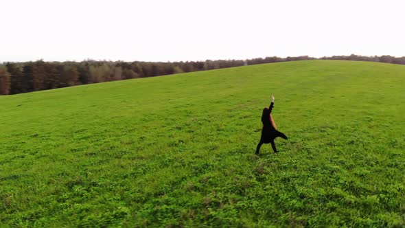 Top View of a Young Girl in a Black Trench Coat Walking Across a Green Field with Her Hands Up to