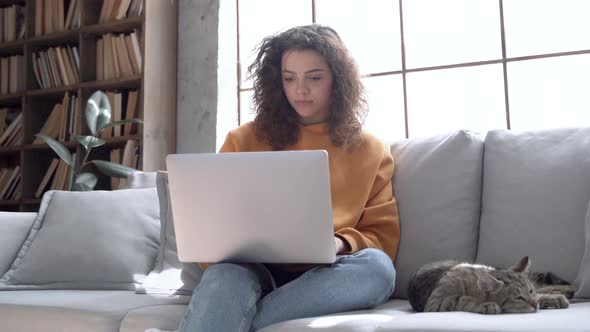 Smiling Hispanic Girl Using Laptop Playing with Cat Sitting on Couch at Home