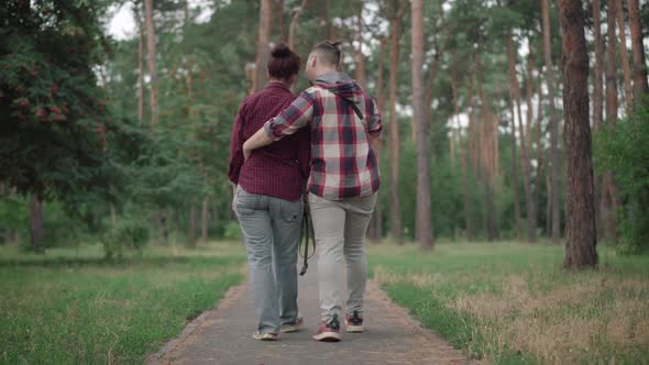 Back View of Loving Caucasian Couple Strolling in Park or Forest with Dog Passing at the Background