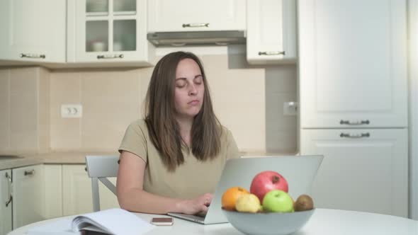 Tired young woman freelancer working on a laptop in the kitchen from home.