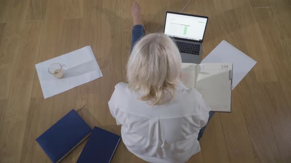 Top View of Caucasian Female Retiree in Casual Clothes Sitting on the Wooden Floor and Using Laptop