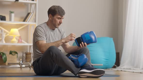 Serious Middle Aged Man Putting on Boxing Gloves Sitting on Exercise Mat