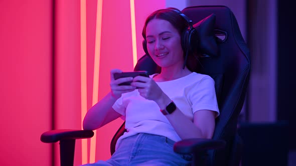 Cute Gamer Girl Sit on a Gaming Chair, Smiling and Playing Mobile Online Game on a Smartphone