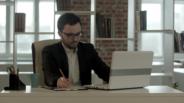 Young Man with Funny Glasses Working on His Notebook, with a Fresh Cup of Coffee Nice and Early in