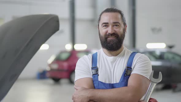 Portrait of a Smiling Bearded Mechanic Posing with a Wrench Standing at an Auto Repair Shop