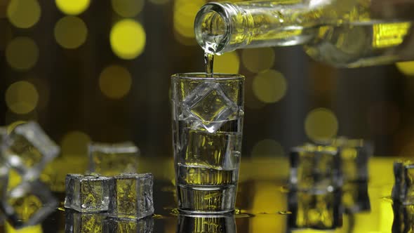 Barman Pour Frozen Vodka From Bottle Into Shot Glass. Ice Cubes Against Shiny Gold Party Background