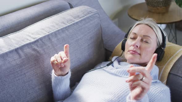 Relaxed senior caucasian woman in living room lying on sofa, wearing headphones
