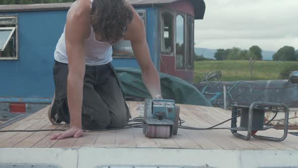 Young carpenter wearing white vest belt sanding cabin roof planking of old wooden boat. MID SHOT.