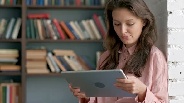 Female Student Sitting Against Bookshelf and Using Laptop in the Library