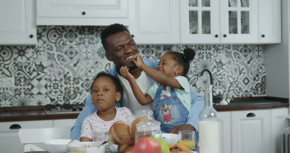 African American which Sitting Near Kitchen Table and Holding on Knees His Two Cute Small Daughters