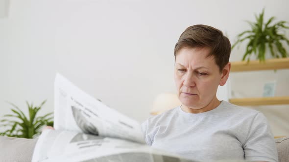 A Closeup View of a Beautiful Mature Woman is Reading a Newspaper Sitting on Sofa at Home in the