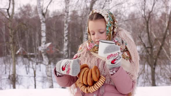 Cute girl in a traditional Russian headscarf is drinking hot tea and eating bagels on winter backgro