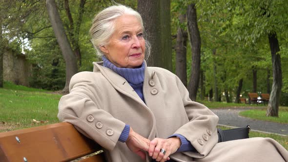 An Elderly Woman Sits on a Bench in a Park and Looks Around, Sad