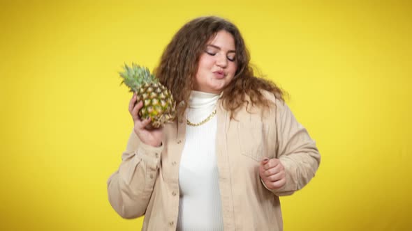 Relaxed Joyful Plussize Woman Dancing with Pineapple at Yellow Background