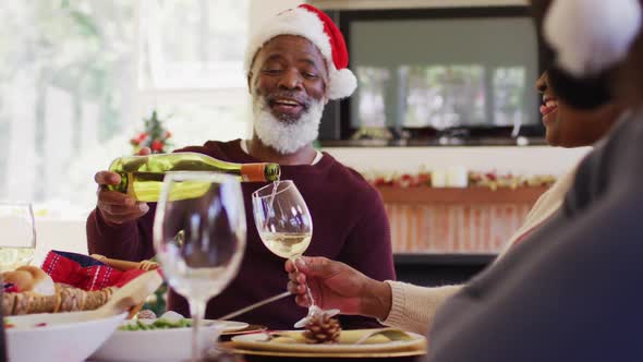 African american senior man in santa hat pouring drink in glass of senior woman while sitting on din