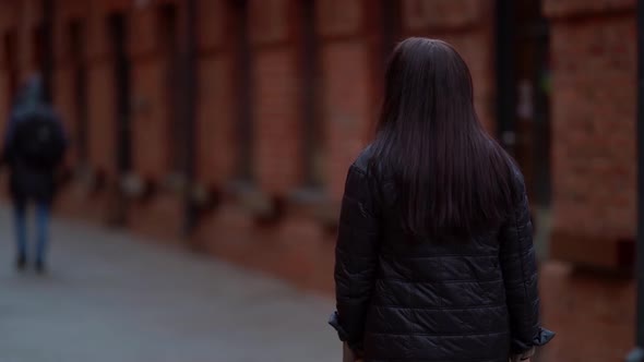 a Woman with Long Hair and a Dark Coat Smiles and Walks Against the Background of a Brick Building