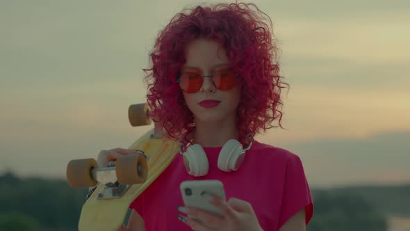 A Pink Curly Teenager is Walking and Using a Smartphone Outdoors
