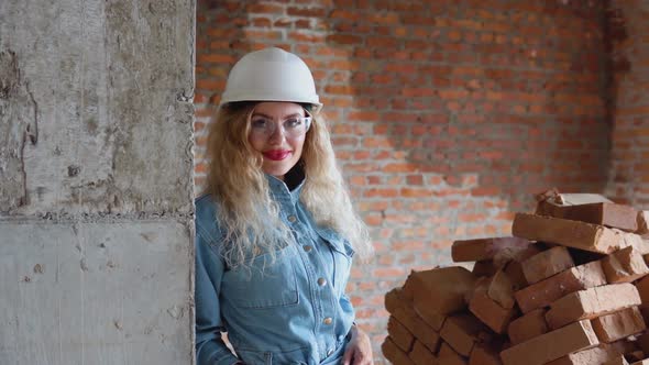 A Young Woman in a Work Helmet and Denim Clothes and Goggles Stands at the Construction Site