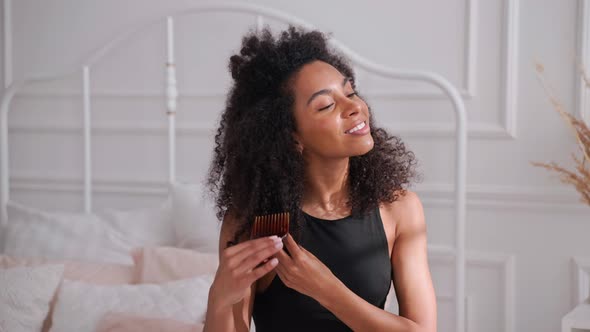 Black Woman Combing Her Afro Hair Sitting on a Bed in Bright White Bedroom
