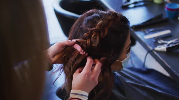 Female hairdresser braiding hair of female customer wearing face mask at hair salon