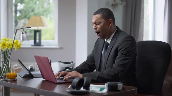 Side View of Concentrated African American Man Typing on Laptop Keyboard and Yawning
