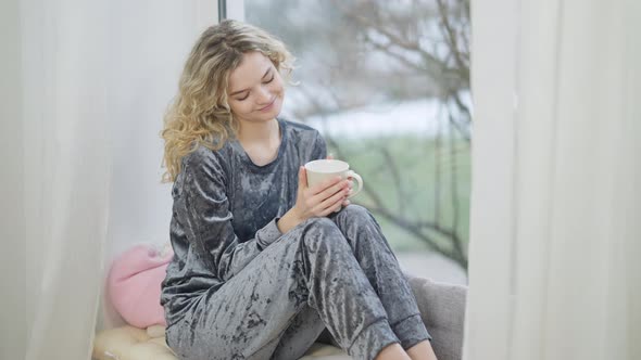 Charming Happy Smiling Woman Sitting on Windowsill Holding Coffee Cup Looking Out the Window