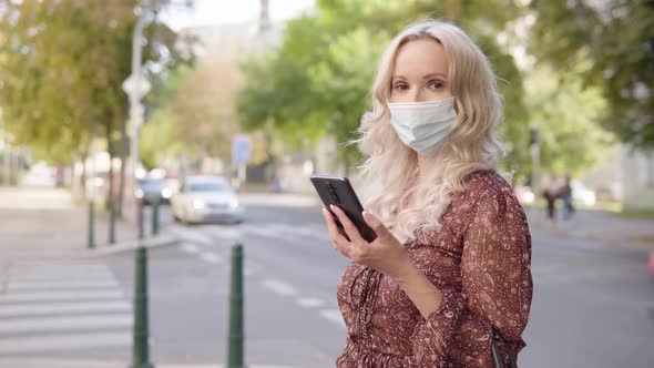 A Middleaged Caucasian Woman in a Face Mask Holds a Smartphone and Looks at Camera in an Urban Area