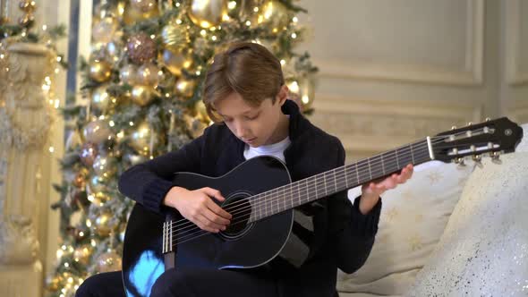 Portrait of Young Guy Playing Guitar Sits on Sofa Against Backdrop of Christmas