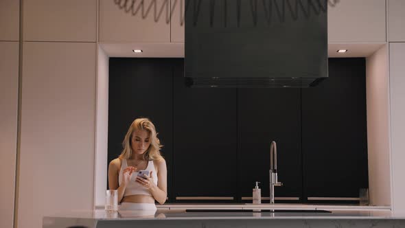 A Young Blonde Woman with a Phone in Her Hands in the Kitchen Chatting